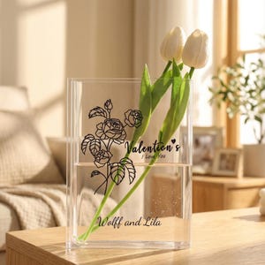 Può includere: Un vaso di vetro trasparente rettangolare con un disegno floreale e il testo "Valentino's I Love You" e "Wolff and Lila". Il vaso contiene due tulipani bianchi con steli e foglie verdi. Il vaso è su una superficie di legno.