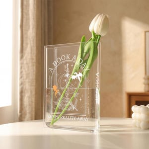 May include: Clear acrylic vase with a white floral design and the text "A BOOK A DAY KEEPS REALITY AWAY." The vase holds a white tulip with green leaves. The vase is on a white table.