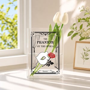 May include: A clear rectangular acrylic vase featuring a black printed design of curtains, the text "THE PHANTOM OF THE OPERA", a white mask, and a red rose. The name "Katniss" is printed at the bottom. Two white tulips are placed inside the vase.