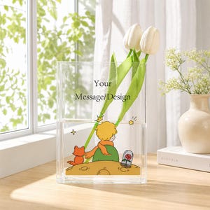 May include: A rectangular clear acrylic vase featuring a printed illustration of a small boy with yellow hair and an orange scarf sitting on a planet with a fox and a rose under a glass dome. The text Your Message/Design is printed in black.