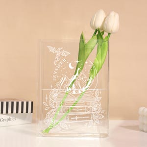 May include: A clear acrylic vase with a white graphic design featuring a dragon, mountains, books, and the name "JENNIFER". The vase holds white tulips. The vase is on a white surface.