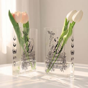 May include: Two clear acrylic vases with black illustrations of mountains, books, and celestial elements. Each vase holds fresh tulips. The word "JENNIFER" is printed vertically on each vase.