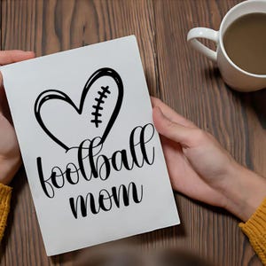 May include: A white greeting card with the words "football mom" in a cursive font. The card features a black heart-shaped design with football stitching. A white mug with a brown liquid sits on a wooden surface.