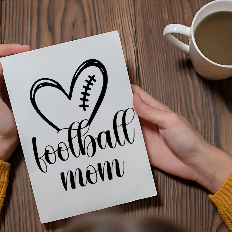 May include: A white greeting card with the words "football mom" in a cursive font. The card features a black heart-shaped design with football stitching. A white mug with a brown liquid sits on a wooden surface.
