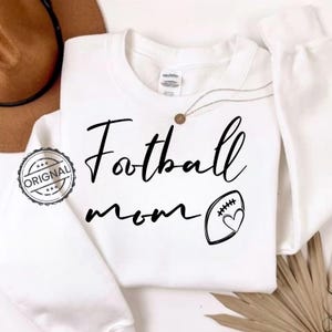 May include: White sweatshirt with the words "Football mom" in black script. A football with a heart is next to the text. A brown hat and a necklace with a pendant are visible in the background.