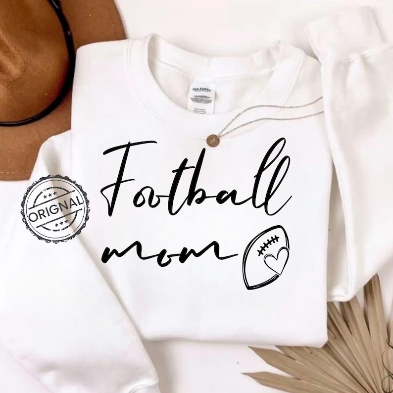 May include: White sweatshirt with the words "Football mom" in black script. A football with a heart is next to the text. A brown hat and a necklace with a pendant are visible in the background.