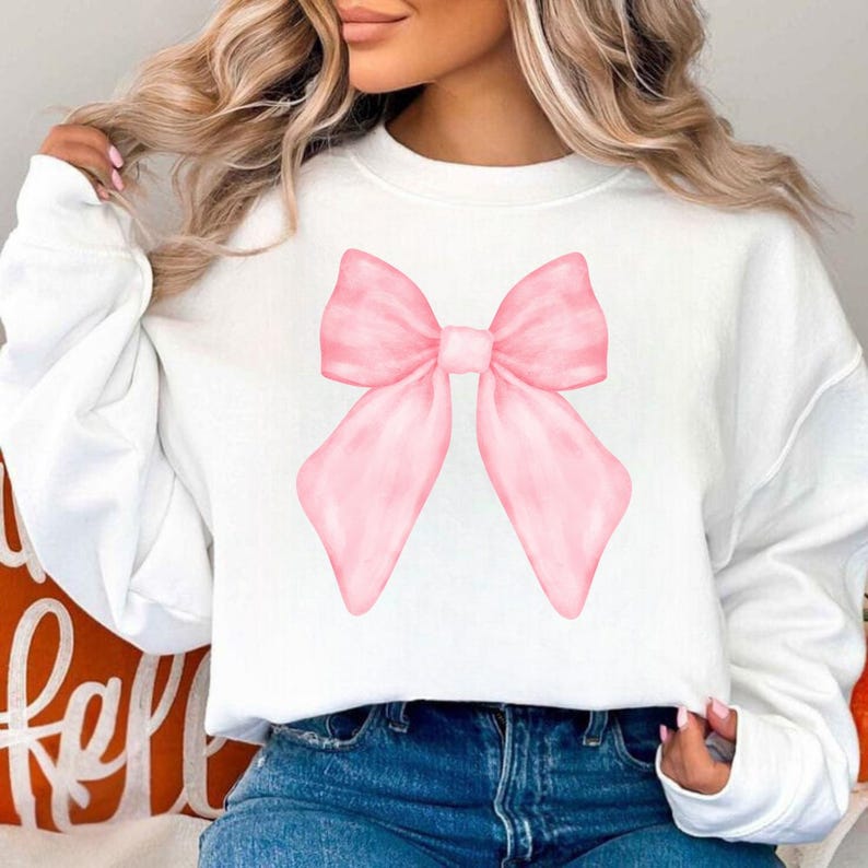 May include: White sweatshirt featuring a large, light pink bow graphic. The bow is centered on the front of the shirt and has a watercolor effect. The sweatshirt has long sleeves and a crew neck.