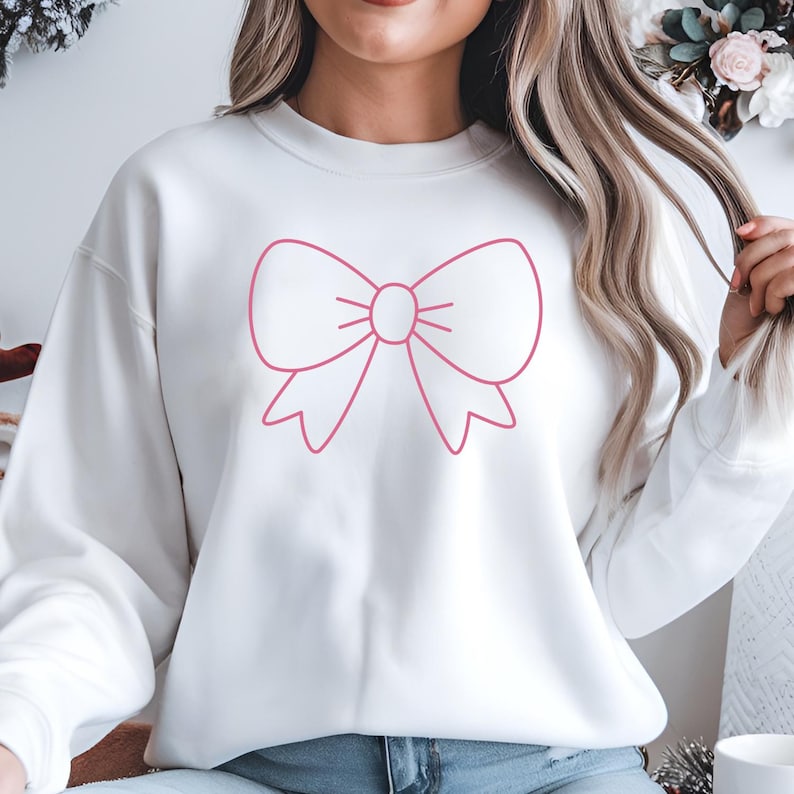 May include: White sweatshirt featuring a large pink bow graphic. The bow is a simple outline design, centered on the front of the garment. The sweatshirt has long sleeves and a crew neck.