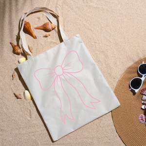 May include: A beige tote bag with a pink bow design. The bag is made of canvas and has a simple handle. The bag is lying on a sandy surface with seashells and a straw hat.