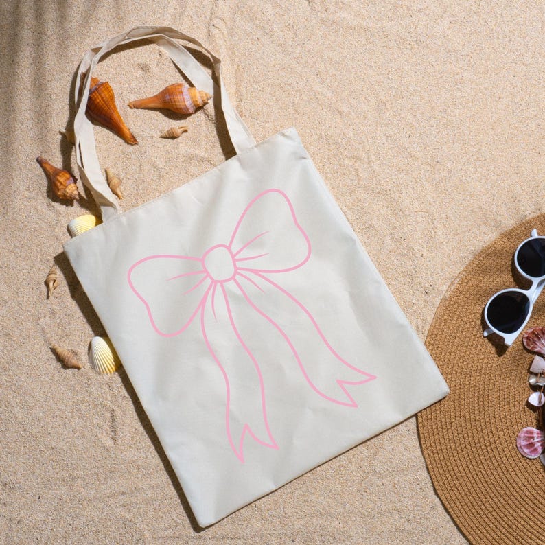 May include: A beige tote bag with a pink bow design. The bag is made of canvas and has a simple handle. The bag is lying on a sandy surface with seashells and a straw hat.