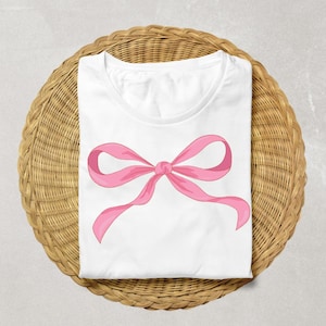 May include: White t-shirt featuring a large pink bow design. The bow is centered on the shirt and has a classic ribbon shape. The shirt is folded neatly on a round, woven wicker placemat.