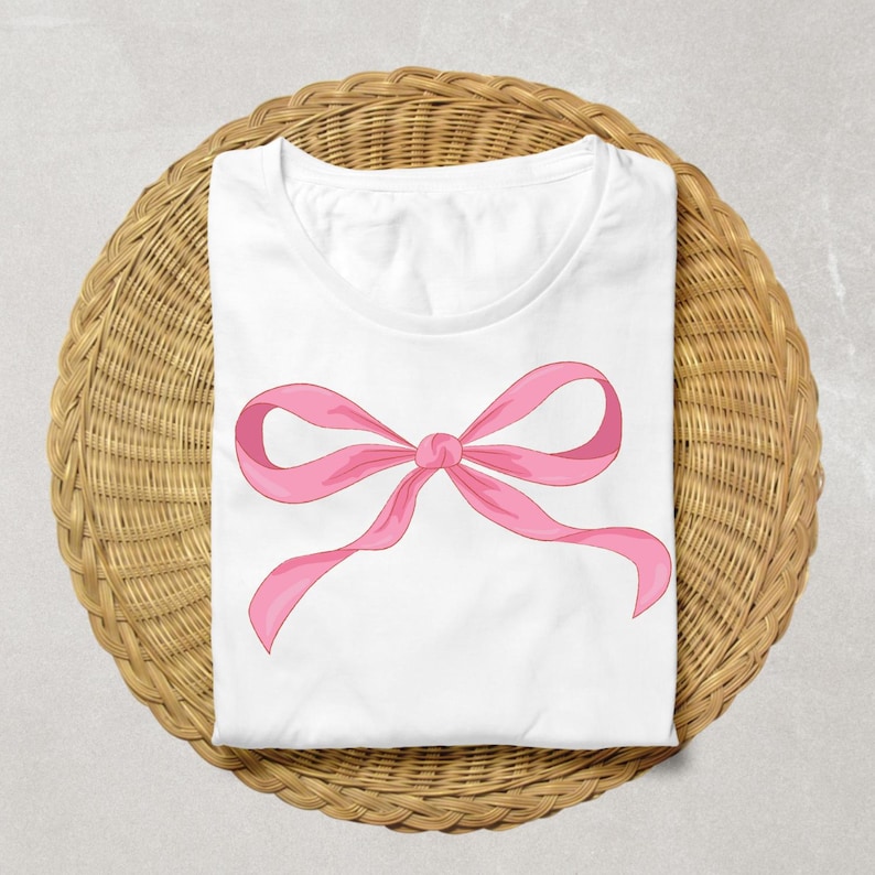 May include: White t-shirt featuring a large pink bow design. The bow is centered on the shirt and has a classic ribbon shape. The shirt is folded neatly on a round, woven wicker placemat.