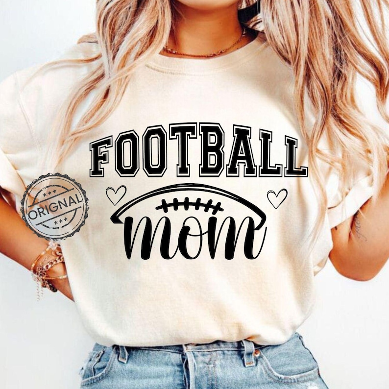 May include: Cream-colored t-shirt with the words "FOOTBALL MOM" in black, with a football graphic and two hearts. The word "ORIGINAL" is stamped on the left side of the shirt.