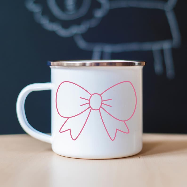 May include: White enamel mug with a pink bow design. The mug has a silver rim and a white handle. The bow is a simple outline, adding a touch of charm to the mug's design. The mug is likely used for beverages.