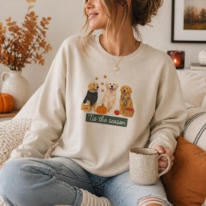 Peut inclure: Sweat-shirt beige avec un motif de trois golden retrievers, des citrouilles et des feuilles d'automne. Le texte "Tis the season" est affiché en dessous des chiens. Le sweat-shirt est de couleur neutre.