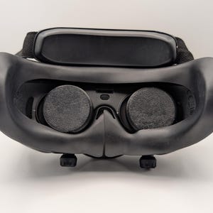 May include: A black virtual reality headset with a head strap and two circular lenses. The headset has a curved design and is made of a matte material. The lenses are dark gray and appear to be textured.