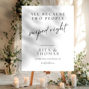 Pode incluir: Uma placa de casamento branca em um cavalete de madeira. O texto diz: "ALL BECAUSE TWO PEOPLE swiped right". Abaixo estão os nomes "RITA & THOMAS" e a data e o local. A placa é cercada por velas e arranjos florais.