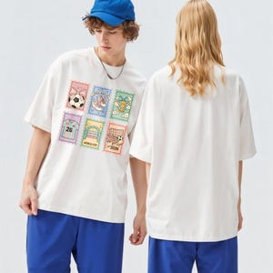 May include: White t-shirt with a colorful graphic of six postage stamp-style images, including a soccer ball, a shoe with a bow, and a soccer goal. The shirt is worn with blue shorts and a blue baseball cap.