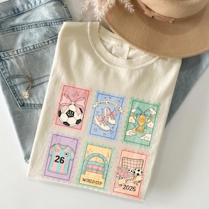 May include: Cream-colored t-shirt featuring colorful postage stamp designs with soccer-themed graphics. Stamps include a soccer ball, soccer shoes, a trophy, a jersey with the number 26, a World Cup arch, and a goal with the text "goal 2026".