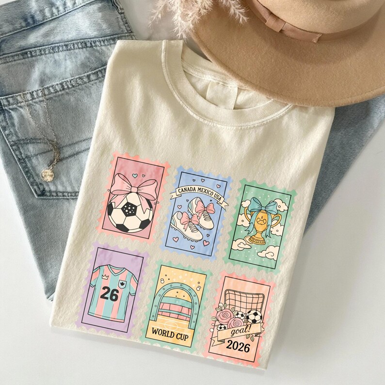 May include: Cream-colored t-shirt featuring colorful postage stamp designs with soccer-themed graphics. Stamps include a soccer ball, soccer shoes, a trophy, a jersey with the number 26, a World Cup arch, and a goal with the text "goal 2026".