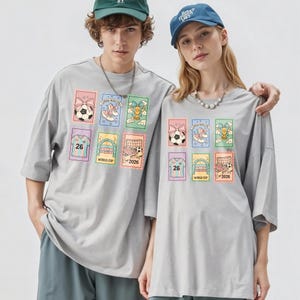 May include: Two people wearing gray t-shirts with colorful soccer-themed graphics. Each shirt features a grid of images including soccer balls, shoes, and a jersey with the number 26. The text "WORLD CUP 2026" is also visible.