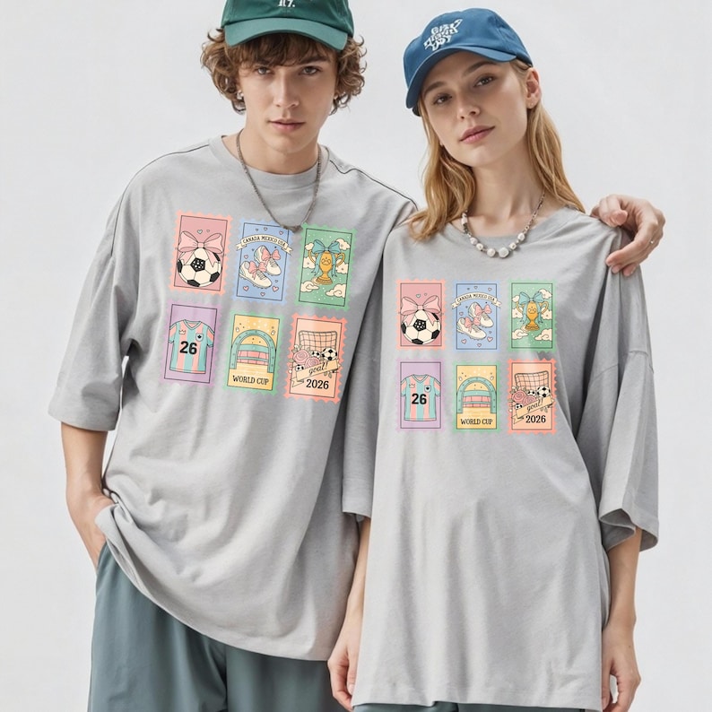 May include: Two people wearing gray t-shirts with colorful soccer-themed graphics. Each shirt features a grid of images including soccer balls, shoes, and a jersey with the number 26. The text "WORLD CUP 2026" is also visible.