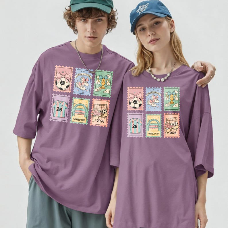 May include: Two people wearing matching purple t-shirts. Each shirt features a colorful stamp design with soccer balls, shoes, and the text "2026". The shirts have short sleeves and a relaxed fit.