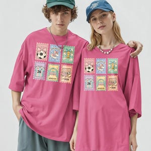 May include: Two people wearing matching magenta t-shirts. Each shirt features a colorful stamp design with soccer balls, shoes, trophies, and the text "WORLD CUP 2026". The shirts have short sleeves and a relaxed fit.
