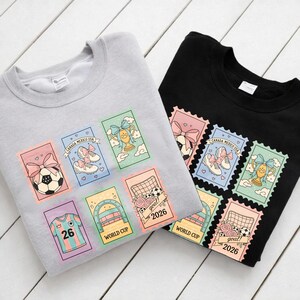 May include: Two sweatshirts, one gray and one black, each adorned with colorful soccer-themed postage stamp designs. The stamps feature soccer balls, cleats, trophies, and the text "CANADA MEXICO USA" and "WORLD CUP 2026".