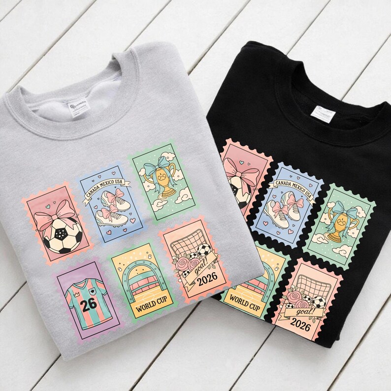 May include: Two sweatshirts, one gray and one black, each adorned with colorful soccer-themed postage stamp designs. The stamps feature soccer balls, cleats, trophies, and the text "CANADA MEXICO USA" and "WORLD CUP 2026".