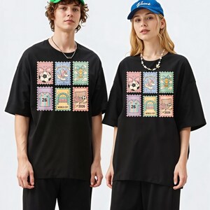 May include: Black t-shirts featuring a colorful stamp design with soccer-themed graphics. The stamps include images of soccer balls, shoes, trophies, jerseys with the number 26, and soccer goals. The text "WORLD CUP 2026" is also visible.