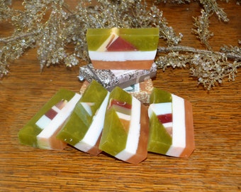 May include: Artisan soap bars with geometric designs. The soaps feature layers of green, white, brown, and red colors. The bars are arranged on a wooden surface with decorative silver branches in the background.