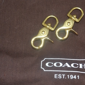 May include: Two gold-toned metal clasps with a hook and loop design. The clasps are on a brown fabric surface with a white COACH logo and the text "EST. 1941".