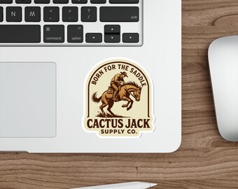 Może przedstawiać: Naklejka z kowbojem na koniu, z napisem "BORN FOR THE SADDLE" i "CACTUS JACK SUPPLY CO." w kolorze brązowym i kremowym. Naklejka znajduje się na białym laptopie. Widoczna jest klawiatura.