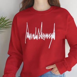 May include: A red sweatshirt with the white text "Ambulance" in a stylized font. The long-sleeved crew neck top is made of a soft material. The sweatshirt is a casual style.