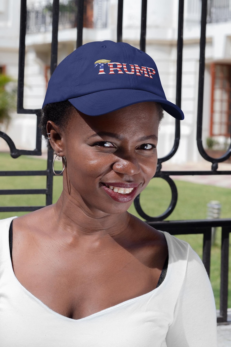 Op de afbeelding: Een marineblauwe baseballpet met het woord "TRUMP" in rode, witte en blauwe letters. De pet is gemaakt van een zacht materiaal en heeft een gebogen klep. De pet wordt door een persoon gedragen.