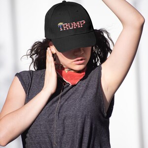 Op de afbeelding: Zwarte baseballpet met het woord "TRUMP" in rode, witte en blauwe letters. De pet heeft een gebogen klep en een klassiek zes-panelen ontwerp. De hoed is een populair politiek accessoire.