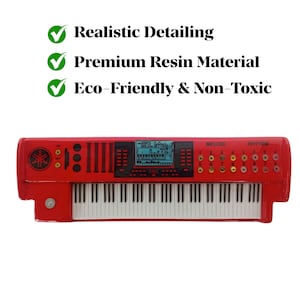 Puede incluir: Un teclado de juguete rojo con teclas blancas y una pantalla digital. El teclado tiene botones y perillas, con el texto "MELODI" y "RHYTHM" visibles. La imagen también incluye el texto "Realistic Detailing", "Premium Resin Material" y "Eco-Friendly & Non-Toxic".