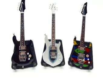 May include: Three miniature electric guitars in black, silver, and a colorful design. Each guitar has a different body shape and is displayed on a black stand. The guitars have detailed fretboards and headstocks.