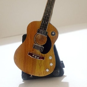 Puede incluir: Una guitarra acústica en miniatura con cuerpo de madera y golpeador negro. La guitarra descansa sobre un soporte negro. El instrumento tiene seis cuerdas y clavijas de afinación plateadas. El cuerpo mide unos 20 cm de largo.