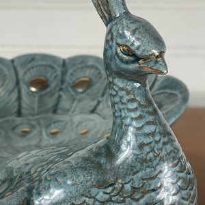 May include: A ceramic peacock-shaped bowl featuring a glossy blue-green glaze with a mottled texture. The peacock head is upright with a crest, and the bowl section displays a fanned tail pattern with gold-colored accents on the eye-like details.