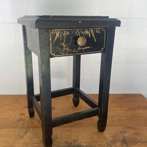 May include: A small black wooden side table featuring a single drawer with a gold-painted bamboo motif and a round brass knob. The table stands on four straight legs connected by a lower support frame, showcasing a dark finish with visible wear.