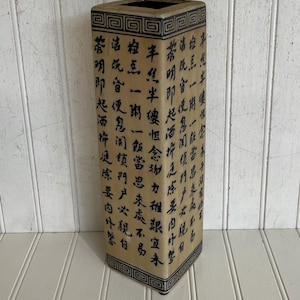 May include: A rectangular beige vase with black Chinese characters. The vase has a dark brown interior and a black and beige geometric design around the top and bottom. The vase is on a white surface.