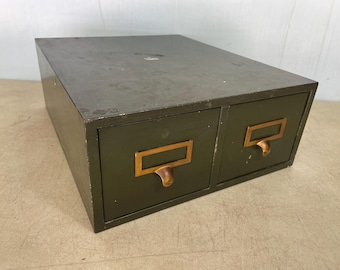 May include: Olive green metal two-drawer storage cabinet with brass-toned label holders and drawer pulls. The cabinet has a rectangular shape and a slightly distressed finish, suggesting vintage or industrial design.