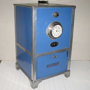 May include: A blue metal Westgate Cooking Company oven with a round clock face and a silver handle. The oven has a silver trim and a silver nameplate.