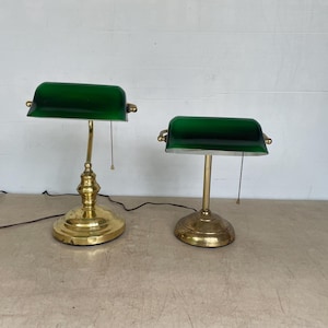 May include: Two banker lamps with polished brass bases and emerald green glass shades. Each lamp features a cylindrical glass shade, a metallic pull chain, and a sturdy circular base. The lamps are connected to black electrical cords.