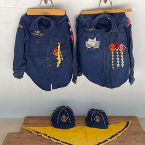 May include: Two navy blue Cub Scout shirts with patches and the text "CUB SCOUTS B.S.A." are displayed on wooden hangers. Below are two navy blue caps with gold emblems and a yellow neckerchief with a blue border.