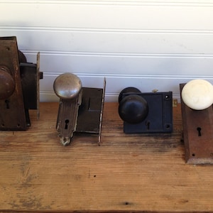 May include: Four antique door knobs with keyholes and plates in various finishes, including black, silver, and brown. The knobs are displayed on a wooden surface.