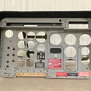 May include: A gray metal aircraft instrument panel frame with a black hood, featuring multiple circular cutouts for gauges, rectangular slots, and several red and black warning placards with white text. The panel has a weathered industrial finish.
