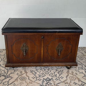 May include: A vintage wooden storage chest with a black leatherette padded top. The chest has two doors with ornate brass hardware and sits on small caster wheels. The wood has a warm, medium-brown tone.